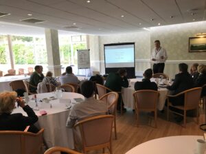 90-day business planning session in progress with the session leader at the front by a whiteboard and tables of delegates facing him, undertaking planning