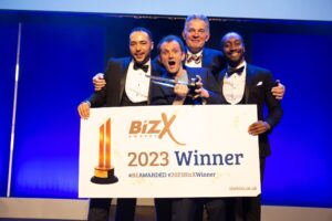 Four men on stage receiving a business awards prize with a banner saying 2023 Winner and BizX Awards. One man is holding a trophy.